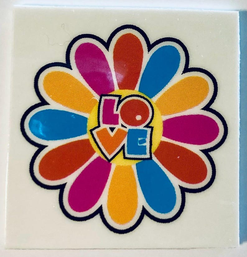 May include: A colorful flower sticker with a black outline. The flower petals are in shades of orange, pink, blue, and yellow. The word "LOVE" is in the center of the flower in a retro font. The sticker is on a white square.