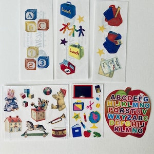May include: A set of colorful stickers featuring various school-themed images, including a lunchbox, a school bus, a clown in a box, a house, a rocking horse, a teddy bear, a chalkboard, a globe, a pencil, a red apple, and a colorful alphabet apple.