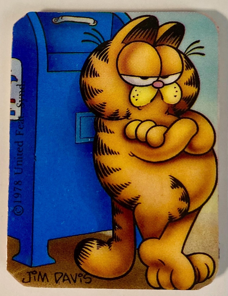 Pu&ograve; includere: Un adesivo vintage di Garfield con il gatto dei cartoni animati appoggiato a una cassetta delle lettere blu. Garfield &egrave; arancione con strisce nere e un'espressione scontrosa. L'adesivo presenta il testo "&copy;1978 United Feature Syndicate, Inc." e "JIM DAVIS".