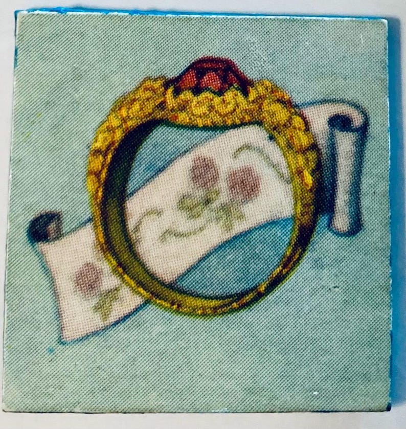 May include: A square image featuring a gold ring with a red gem on top, encircling a white scroll with pink floral designs. The background is a light blue, and the ring has intricate detailing.