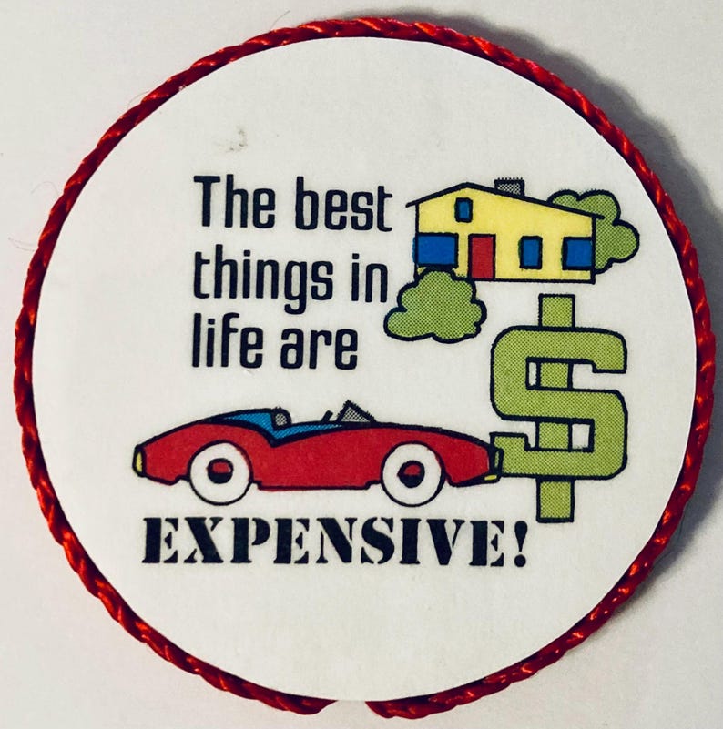 May include: A round white pinback button with a red braided border. It features a yellow house, a red convertible car, and a large green dollar sign. The text reads: The best things in life are EXPENSIVE! The design uses black, red, yellow, and green.