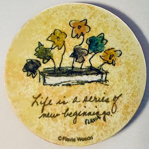 May include: Round coaster with a floral design and the text "Life is a series of new beginnings!" in black script. The coaster features a watercolor-style illustration of colorful flowers in a planter.