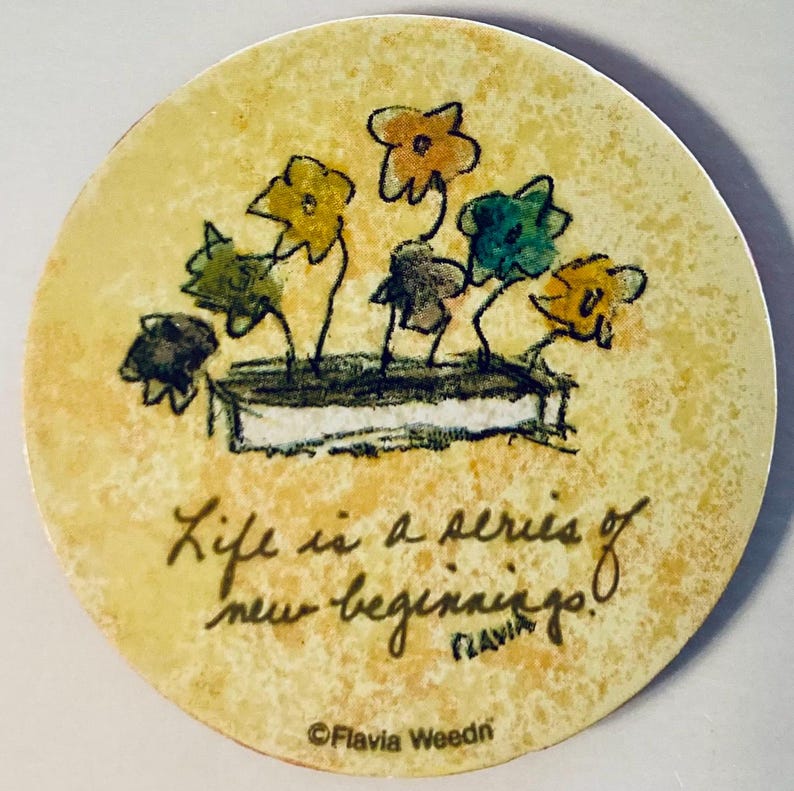 May include: Round coaster with a floral design and the text "Life is a series of new beginnings!" in black script. The coaster features a watercolor-style illustration of colorful flowers in a planter.