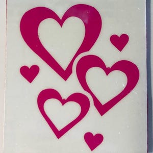 May include: A white square tile featuring a design of pink hearts. The design includes three large hearts and three smaller hearts. The hearts are a vibrant shade of pink, creating a contrasting design against the white background.