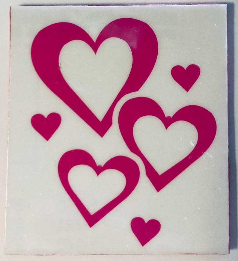 May include: A white square tile featuring a design of pink hearts. The design includes three large hearts and three smaller hearts. The hearts are a vibrant shade of pink, creating a contrasting design against the white background.