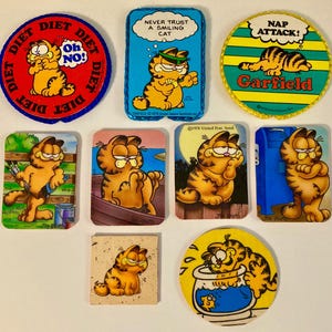 May include: A collection of vintage Garfield the cat collectibles. The items include circular and rectangular patches and cards with Garfield illustrations and phrases like "Diet," "Never Trust a Smiling Cat," and "Nap Attack!"