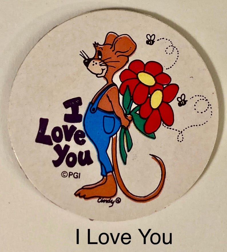 May include: A round, off-white button with a cartoon mouse wearing blue overalls and holding red flowers with yellow centers. The text "I Love You" is in purple, with two small bees and a dotted line design.
