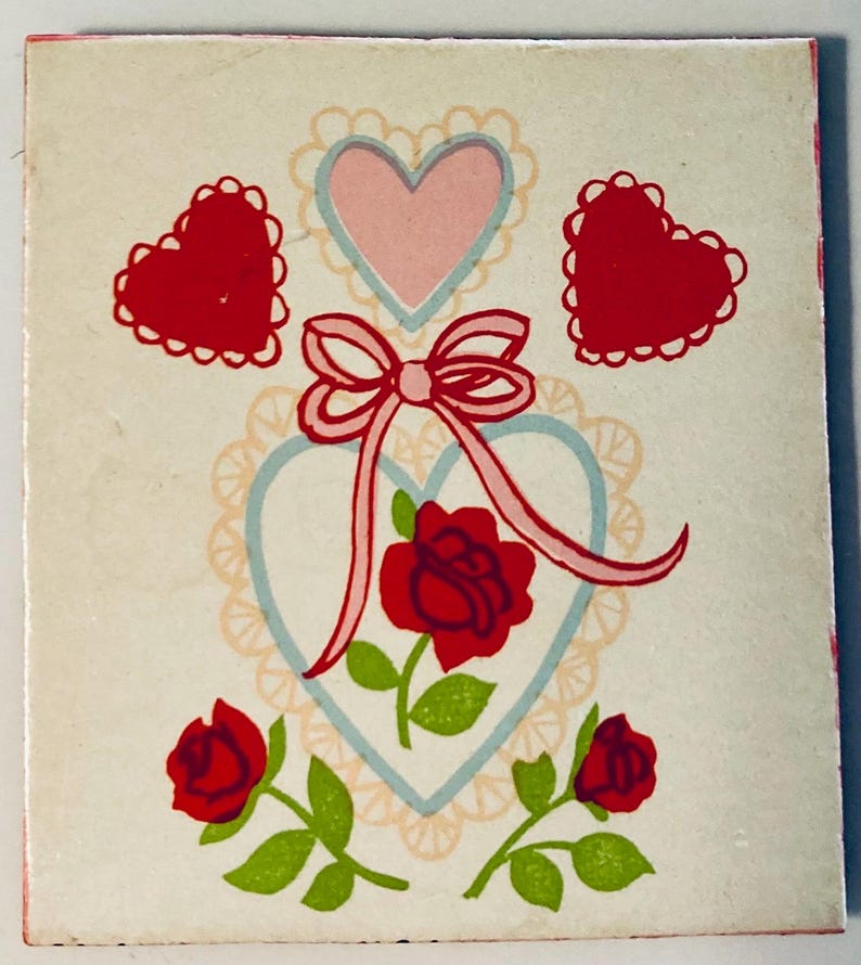 May include: A vintage greeting card with a white background featuring red hearts, roses, and a pink bow. The card has a light blue heart outline with a pink heart inside. Green leaves and stems accent the roses.