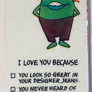 May include: A rectangular card with a cartoon character and text. The green character has orange hair and brown pants. Text reads: "I LOVE YOU BECAUSE...YOU LOOK SO GREAT IN YOUR DESIGNER JEANS...YOU NEVER HEARD OF DESIGNER JEANS...YOU HAVE A JOB."