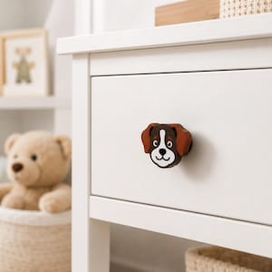 Könnte beinhalten: Ein Schubladenknopf in Form eines Hundekopfes mit braunen Ohren, weißer Schnauze und dunkelbraunem Gesicht. Der Knopf ist an der Vorderseite einer weißen Holzschublade befestigt und zeigt ein Cartoon-Design mit schwarzen Augen, einer schwarzen Nase und einem Mund.