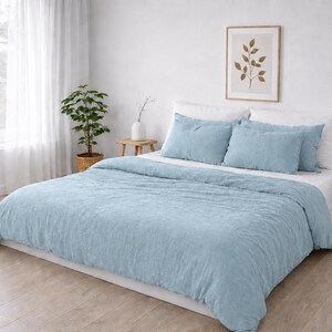 May include: A light blue duvet cover set featuring a textured fabric finish on the bed. The set includes two matching rectangular pillow shams and a large duvet cover that drapes over the sides of the bed, creating a soft and uniform appearance.