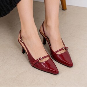 May include: A pair of dark red patent leather slingback heels with a pointed toe. Each shoe features a thin strap across the vamp with a small gold-toned metal buckle and an adjustable slingback strap with a matching gold-toned buckle closure.