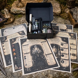 May include: A black gift box containing a small glass vial of crystals, a scroll, and various items, surrounded by several printed informational cards titled Sacred Crystalarium, Moon-Aligned Crystals, and Crystal Cleansing Methods.