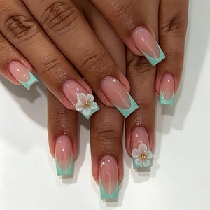 Puede incluir: Uñas largas de forma cuadrada con estilo de manicura francesa con una punta de color verde menta suave sobre una base nude. Dos uñas de acento están adornadas con un adorno floral blanco tridimensional con una pequeña cuenta dorada.