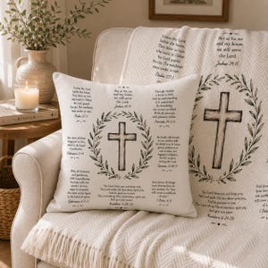 May include: A cream-coloured throw pillow and matching blanket featuring a printed design of a central cross surrounded by a leafy wreath. The fabric displays various Bible verses in black text, including Joshua 24:15, Psalm 127:1, and Proverbs 24:3-4.