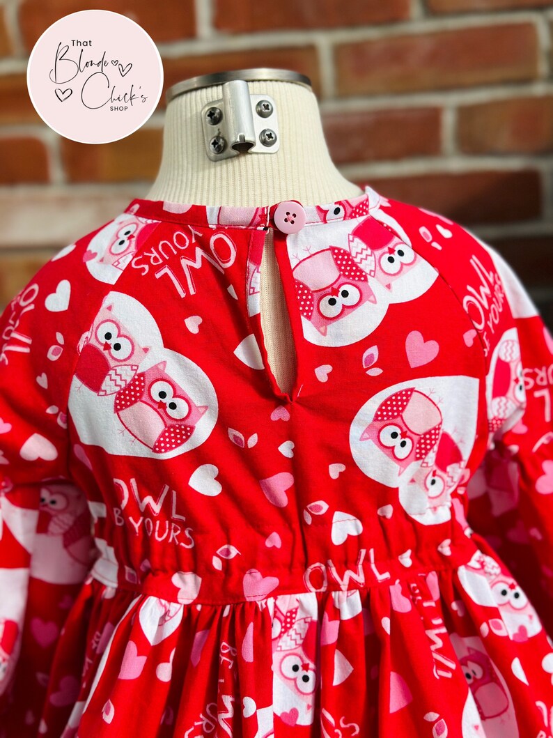 May include: Red dress with a white and pink owl print. The dress has long sleeves and a button closure in the back. The dress has the text "Owl & Yours" repeated on the fabric.