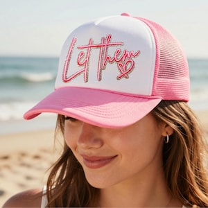 May include: A white and pink trucker hat featuring a pink script graphic that reads Let Them with a small heart icon. The cap has a solid white front panel, a pink brim, and a pink mesh back panel for ventilation, designed with a structured crown.