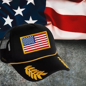 May include: A black trucker cap featuring a rectangular embroidered patch of the United States flag with a gold border. The cap includes a gold cord trim across the base of the crown and gold embroidered laurel leaf designs on the peak.