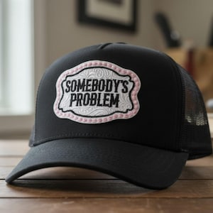 Peut inclure: Une casquette trucker noire avec un écusson blanc à bordure décorative portant l'inscription SOMEBODY'S PROBLEM en lettres noires. L'écusson présente un bord cousu rose clair et un motif floral en relief sur le fond blanc.