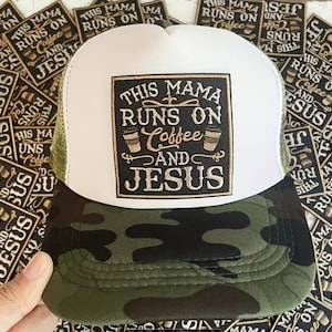 May include: A trucker hat with a white front panel, olive green mesh back, and a camouflage brim. A square patch on the front features the text "THIS MAMA RUNS ON COFFEE AND JESUS" in gold and black, accompanied by two coffee cup illustrations.