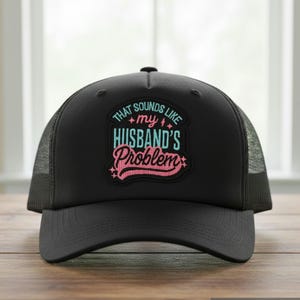 May include: A black trucker hat featuring a central patch with the text "THAT SOUNDS LIKE my HUSBAND'S Problem" in light blue and pink lettering, accented with small star and lightning bolt symbols on a dark background.