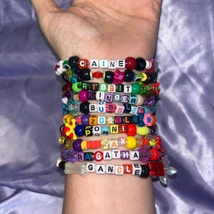 Puede incluir: Una pila de pulseras de cuentas de plástico multicolor con cuentas de letras que deletrean CAINE, RABBIT, KINGER, BUBBLE, ZOOBLE, POMNI, JAX, RAGATHA y GANGLE. Las pulseras incluyen varias formas, tamaños y colores de cuentas y dijes.