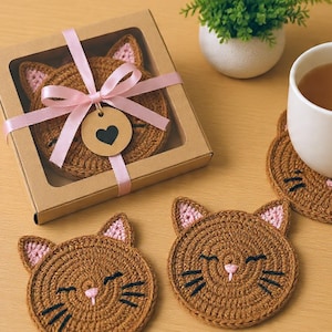 Könnte beinhalten: Ein Set aus braunen, gehäkelten Untersetzern in Katzenform mit rosa Innenohren sowie schwarzen gestickten Augen und Schnurrhaaren. Ein Untersetzer befindet sich in einer braunen Pappschachtel mit rosa Schleife und einem runden Herz-Anhänger.