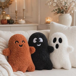 May include: Three plush ghost-shaped pillows made of textured, fuzzy fabric. One pillow is burnt orange with a smiling face, one is black with a smiling face and white eyes, and one is white with a surprised face featuring black eyes and mouth.