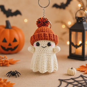 May include: A crochet ghost keychain featuring a cream-colored body with black bead eyes, a small smile, and pink cheeks. The ghost wears a textured orange beanie topped with a multi-colored pom-pom, attached to a silver-toned metal keyring.