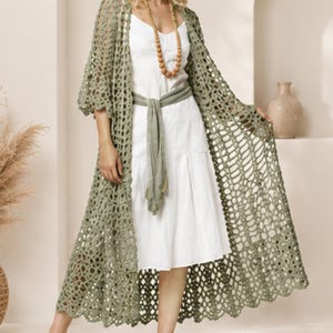 May include: A long, sage green open-front cardigan featuring an intricate, repeating crochet lace pattern with wide sleeves. It is layered over a simple white midi dress cinched at the waist with a matching sage green fabric sash belt.