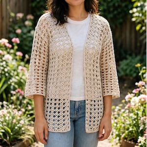 May include: A beige open-front cardigan featuring a repeating geometric crochet pattern with three-quarter length sleeves. The garment is worn over a plain white t-shirt and light blue denim jeans, showcasing a lightweight, textured knit construction.
