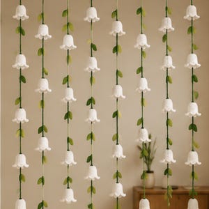 May include: A decorative curtain featuring eight vertical strands suspended from a horizontal rod. Each strand consists of a green cord adorned with small green fabric leaves and multiple white crocheted bell-shaped flowers hanging downwards.