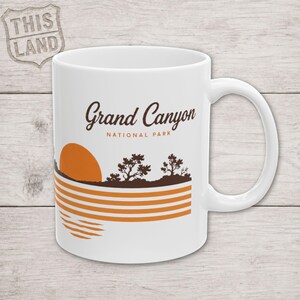 Peut inclure: Un mug en céramique blanc orné d'un graphisme du parc national du Grand Canyon. Le motif comprend du texte marron, un soleil orange, des silhouettes d'arbres marron foncé et des rayures horizontales orange représentant un paysage au coucher du soleil.