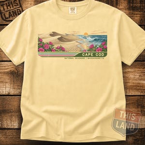 May include: A pale yellow t-shirt featuring a graphic illustration of the Cape Cod National Seashore in Massachusetts. The design depicts sand dunes, ocean waves, a sunset, and pink flowers, with the text CAPE COD NATIONAL SEASHORE MASSACHUSETTS.