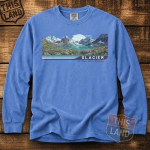 Puede incluir: Una camiseta de manga larga en color azul bígaro con un estampado gráfico de un paisaje montañoso con un lago y una luna grande. El texto dice GLACIER NATIONAL PARK | MONTANA. La camiseta tiene cuello redondo y una etiqueta de Comfort Colors.
