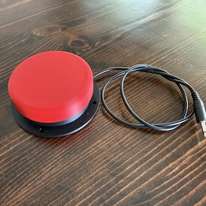 May include: A circular red push button switch mounted on a black circular base with a black cord and a 3.5mm audio jack connector. The device features a smooth, matte red top surface and a black plastic housing with visible mounting holes.