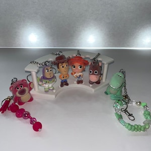 May include: A collection of six character keychains featuring a pink bear, a space ranger, a cowboy, a cowgirl, a horse, and a green dinosaur. The figures are arranged on a white curved display stand, with beaded attachments on the outer keychains.
