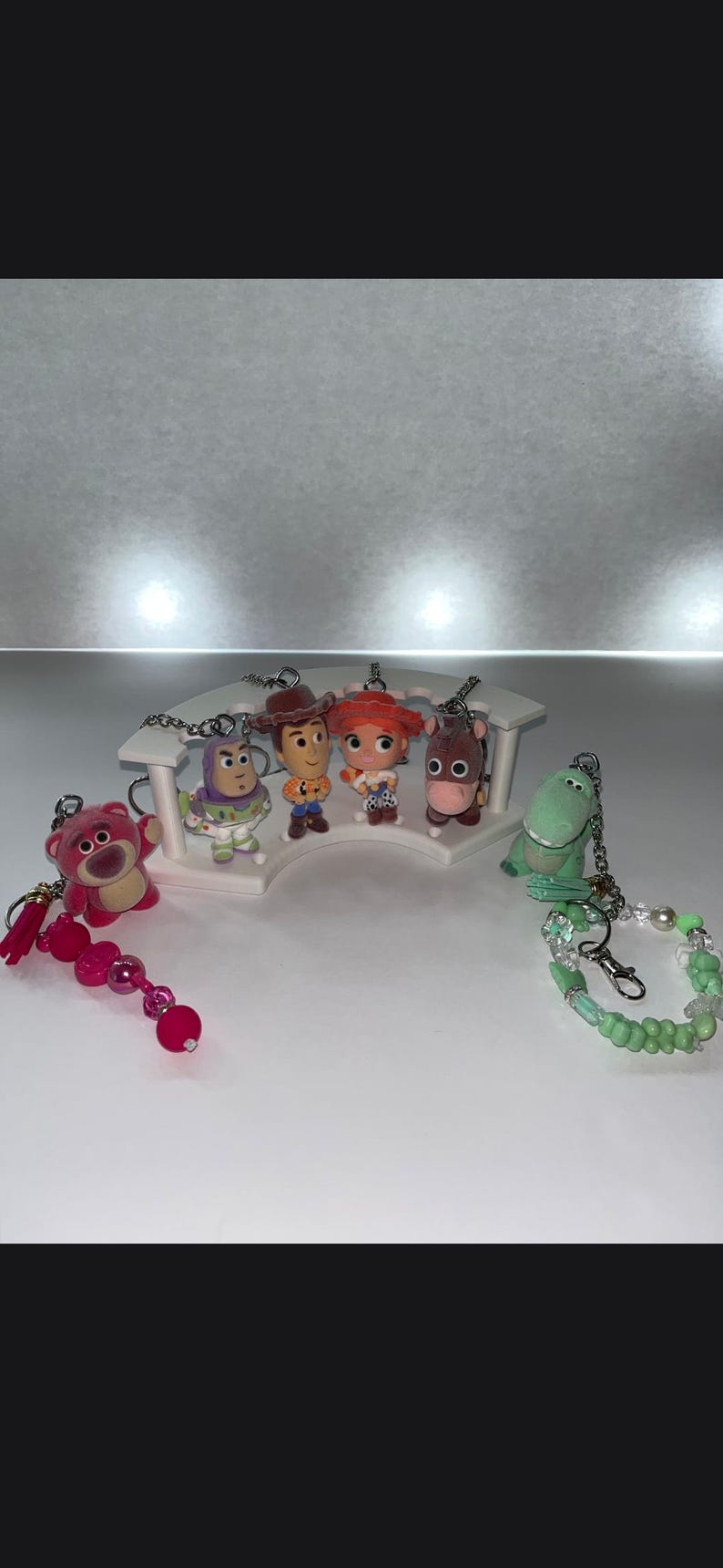 May include: A collection of six character keychains featuring a pink bear, a space ranger, a cowboy, a cowgirl, a horse, and a green dinosaur. The figures are arranged on a white curved display stand, with beaded attachments on the outer keychains.