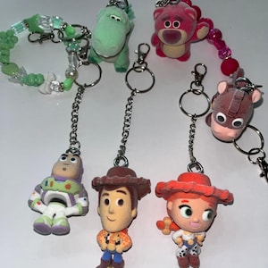 May include: A collection of six character keychains featuring a green dinosaur, a pink bear, a brown horse, a space ranger, a cowboy, and a cowgirl. The set includes a green beaded bracelet attachment and silver metal chains with rings.
