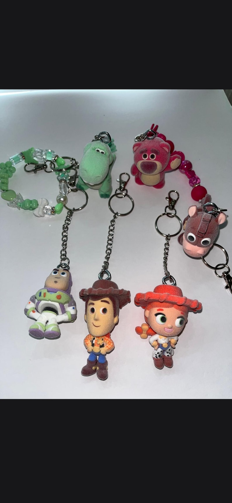 May include: A collection of six character keychains featuring a green dinosaur, a pink bear, a brown horse, a space ranger, a cowboy, and a cowgirl. The set includes a green beaded bracelet attachment and silver metal chains with rings.
