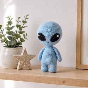 May include: A light blue crochet alien doll with a large rounded head, black oval eyes featuring white highlights, and a small embroidered mouth. The figure has a compact body with short arms and legs, standing upright with a textured yarn finish.