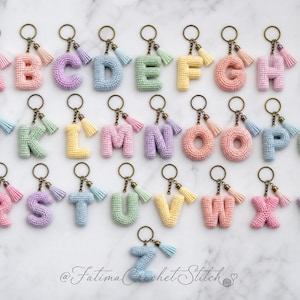 Può includere: Una collezione di ventisei portachiavi con lettere all'uncinetto, ognuno caratterizzato da una lettera in filato color pastello attaccata a un anello in metallo color bronzo e una piccola nappa abbinata. Il testo Fatima Crochet Stitch è visibile in basso.