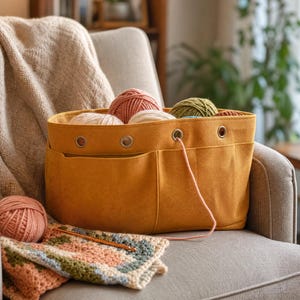 May include: A mustard yellow felt yarn storage bag with multiple exterior pockets and metal grommets along the top edge. The bag is filled with several balls of yarn in shades of pink, cream, and green, with a crochet project and hook nearby.