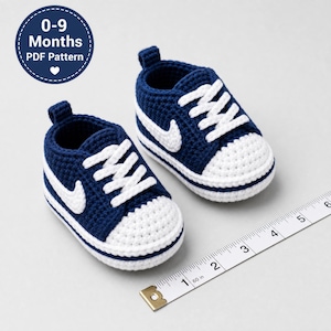 Könnte beinhalten: Ein Paar dunkelblaue und weiße gehäkelte Babysneaker mit weißen Schnürsenkeln und einem seitlichen Swoosh-Design. Ein Maßband zeigt, dass die Schuhe etwa 10 cm lang sind. Ein kreisförmiges Abzeichen zeigt den Text 0-9 Months PDF Pattern.