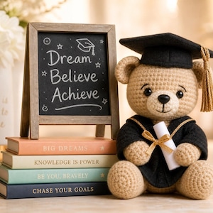 May include: A tan crocheted teddy bear wearing a black graduation cap with a gold tassel and a black gown, holding a white diploma. Next to it is a small chalkboard with the text Dream Believe Achieve and a stack of four books with titles.