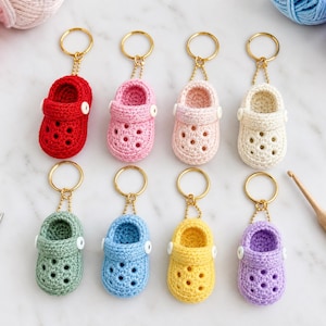 May include: Eight miniature crochet clogs arranged in two rows, each featuring a gold-toned metal keyring, a small white button detail on the side, and perforated holes. The clogs are in red, pink, light pink, cream, sage green, blue, yellow, and purple.