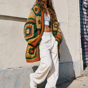 Puede incluir: Un cárdigan de ganchillo de manga larga con un patrón de cuadrados de la abuela multicolor en tonos verde bosque, amarillo mostaza y naranja quemado. La prenda se lleva sobre una camiseta corta blanca, con pantalones cargo blancos y zapatillas.