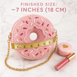 Pode incluir: Uma bolsa de crochê rosa em formato de donut com granulados brancos, vermelhos e beges, com alça de corrente dourada. Uma fita métrica mostra um diâmetro de 18 cm. Texto: FINISHED SIZE: ~7 INCHES (18 CM). Inclui um compacto rosa e um brilho labial.