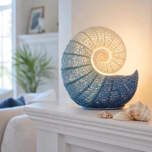 May include: A nautilus shell-shaped table lamp featuring a gradient crochet texture that transitions from a warm cream color at the center to a deep blue at the outer edge, emitting a soft internal glow through the woven material.