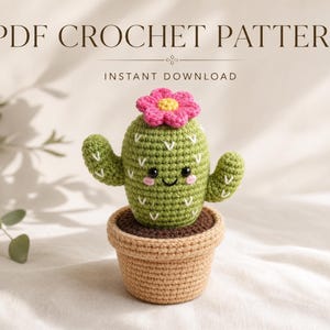 May include: A PDF crochet pattern for a green cactus with white V-shaped embroidery, a pink flower with a yellow center, and a smiling face, sitting in a tan crocheted pot. The text reads PDF CROCHET PATTERN and INSTANT DOWNLOAD.